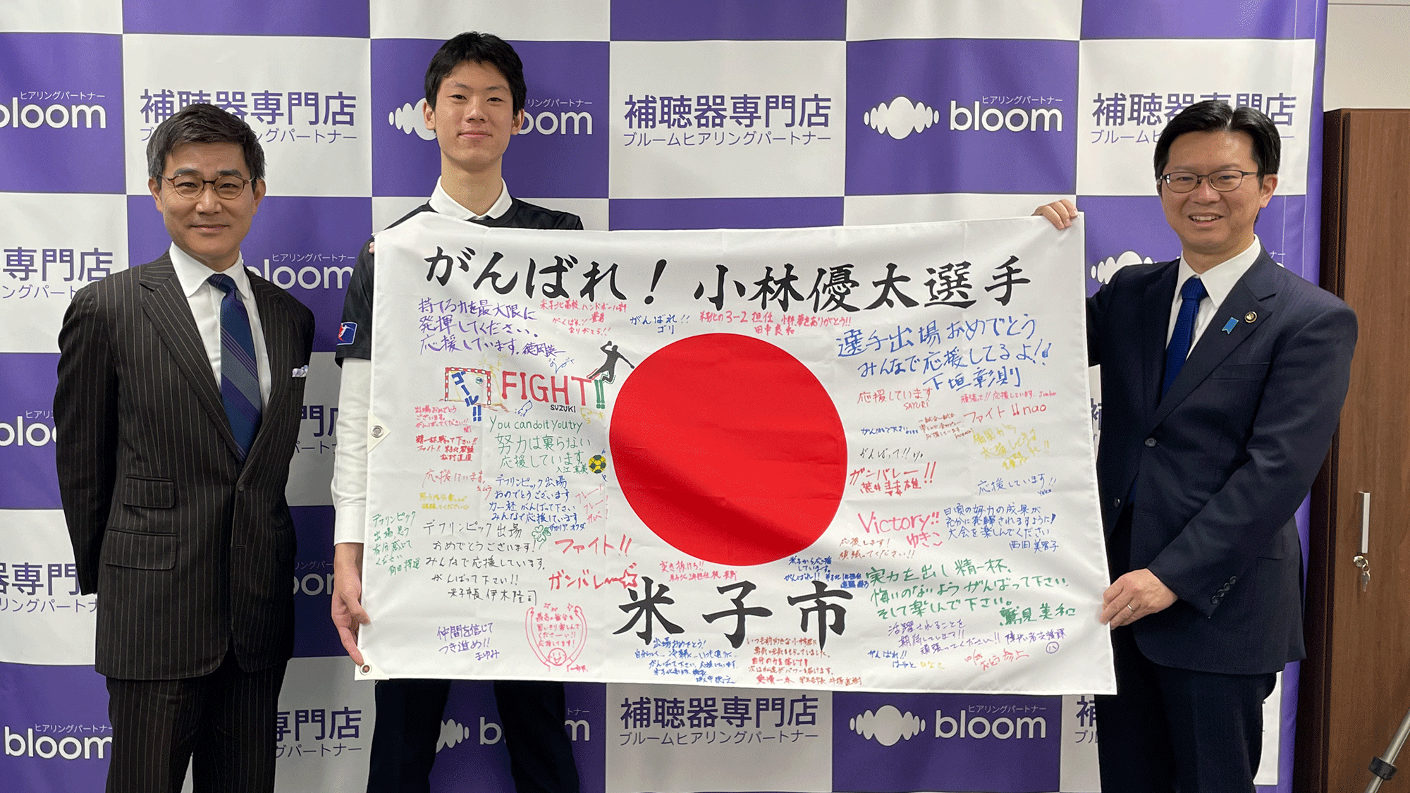 小林選手、伊木市長、松崎社長が国旗を広げて見せる様子
