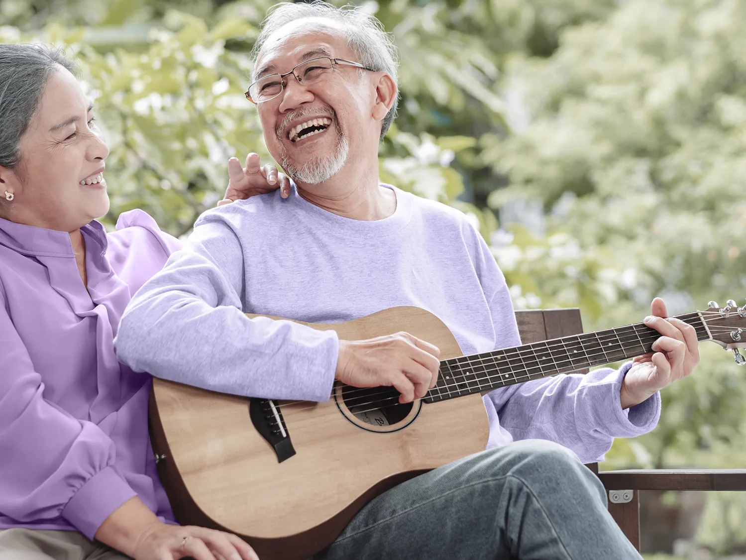 Happy senior couple wearing hearing aids and playing guitar outside house near backyard garden