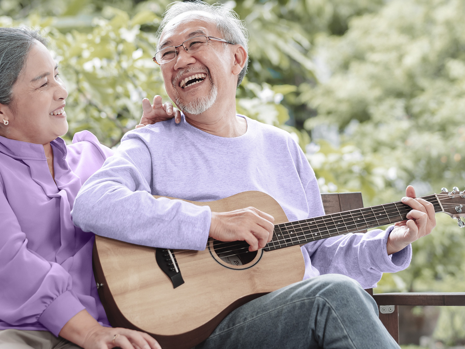 Happy senior couple wearing hearing aids and playing guitar outside house near backyard garden