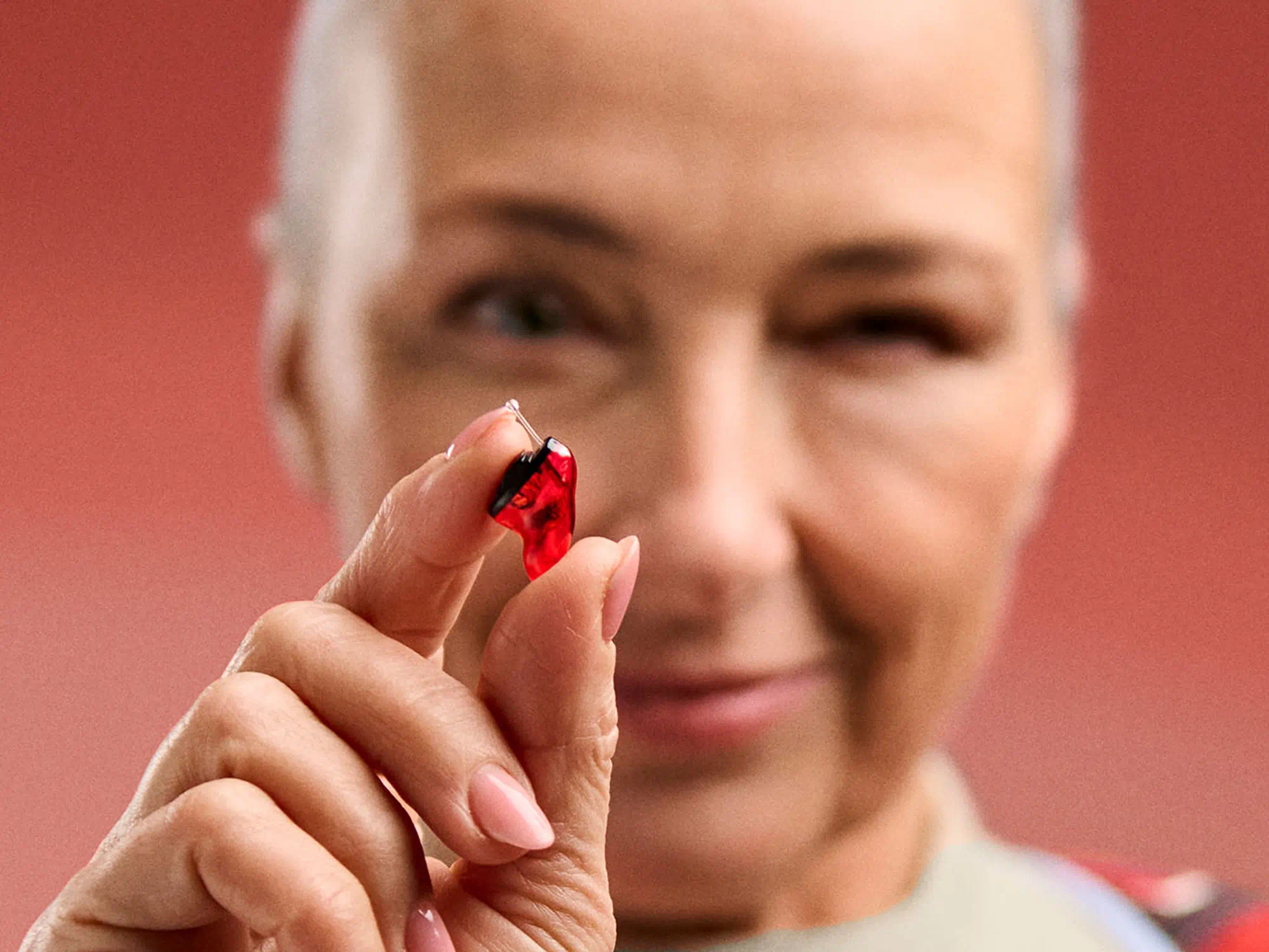 Older adult with hearing-loss holding a small hearing-aid against a red background, highlighting ease of use and comfort.
