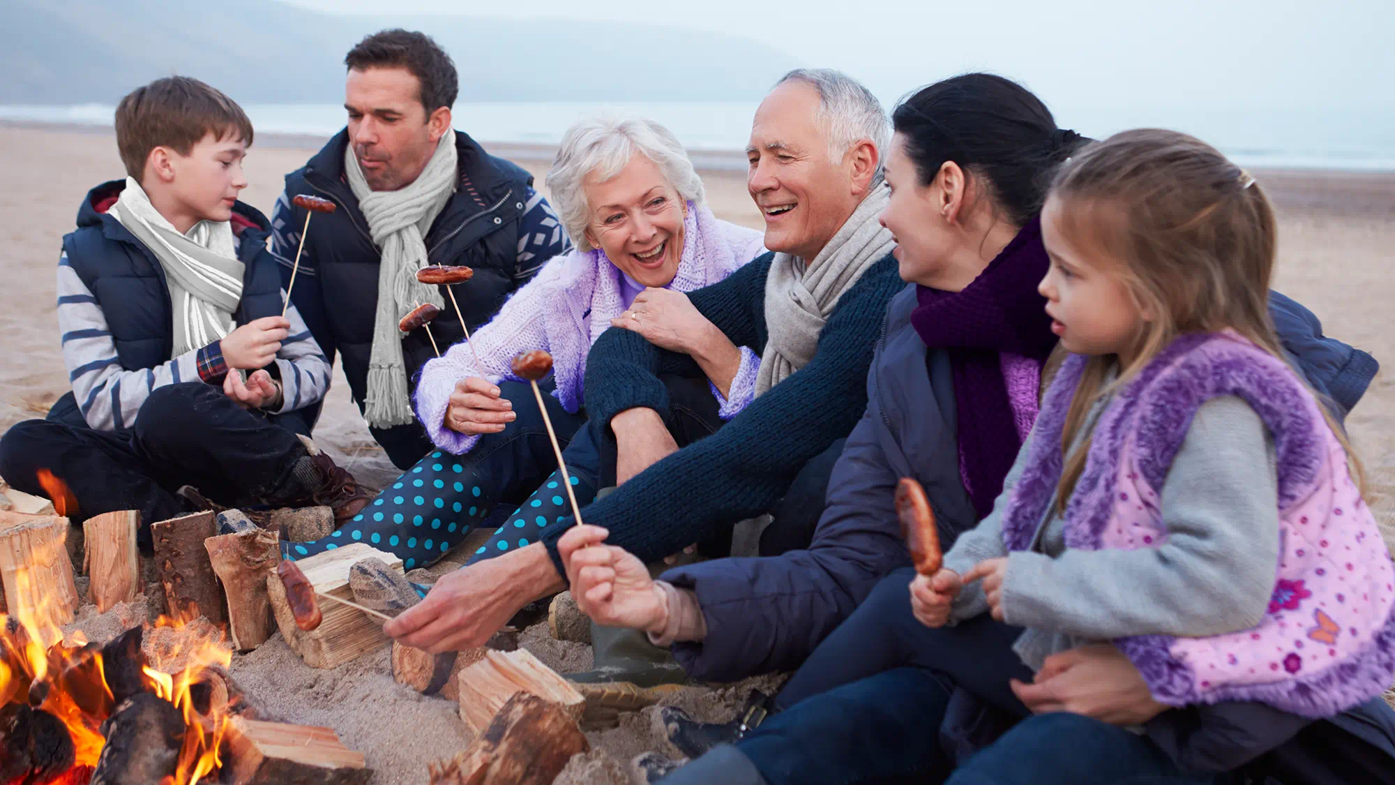 Family in winter gear around a beach campfire, illustrating cold-weather hearing aid care.